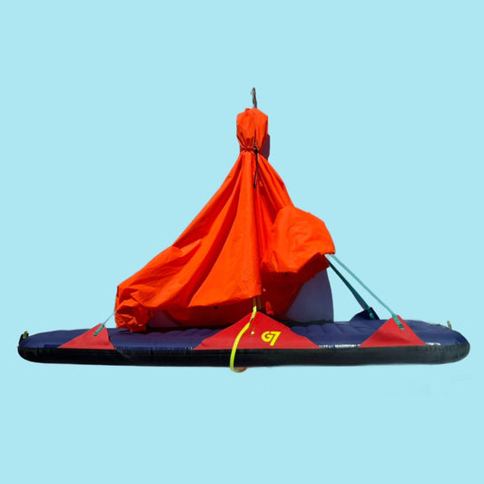 A red and blue portaledge climbing accessory with a waterproof cover on top, displayed against a clear blue sky.