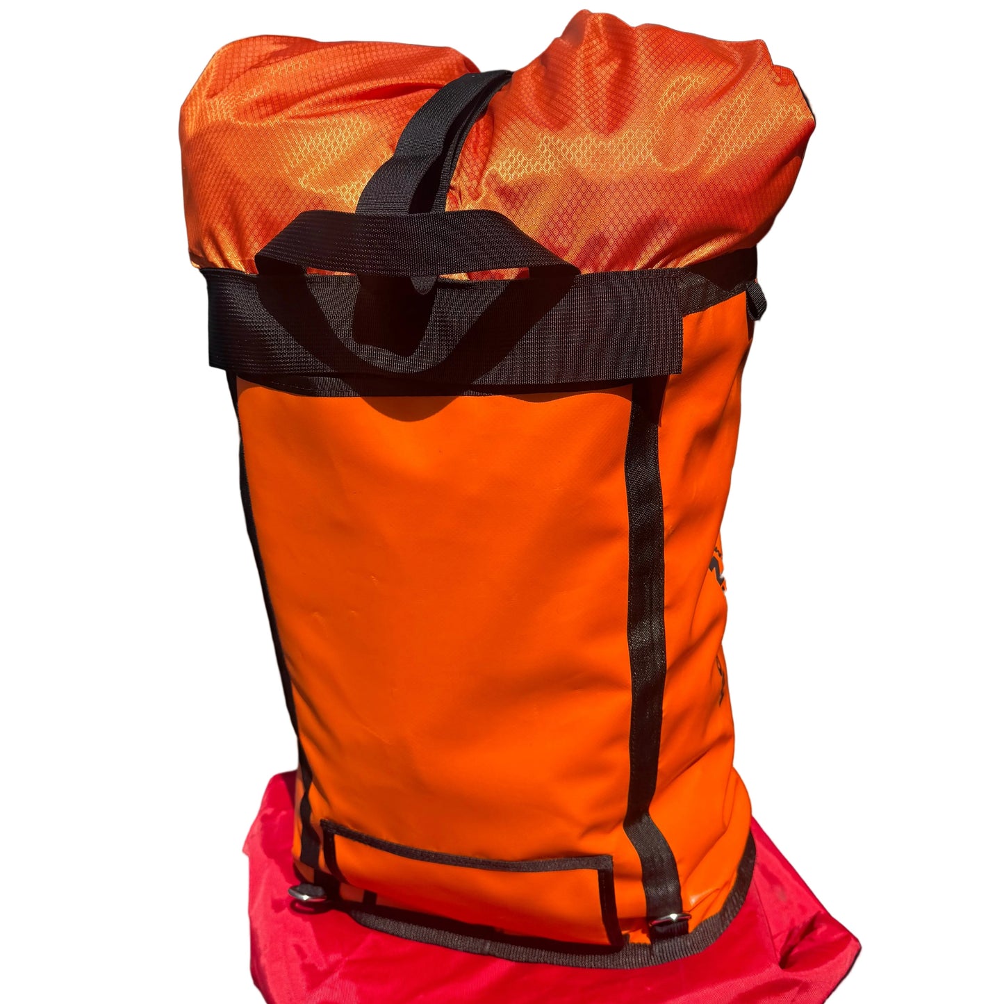 An orange waterproof stuff sack with black straps and trim, resting on top of a red bag against a dark brick wall.