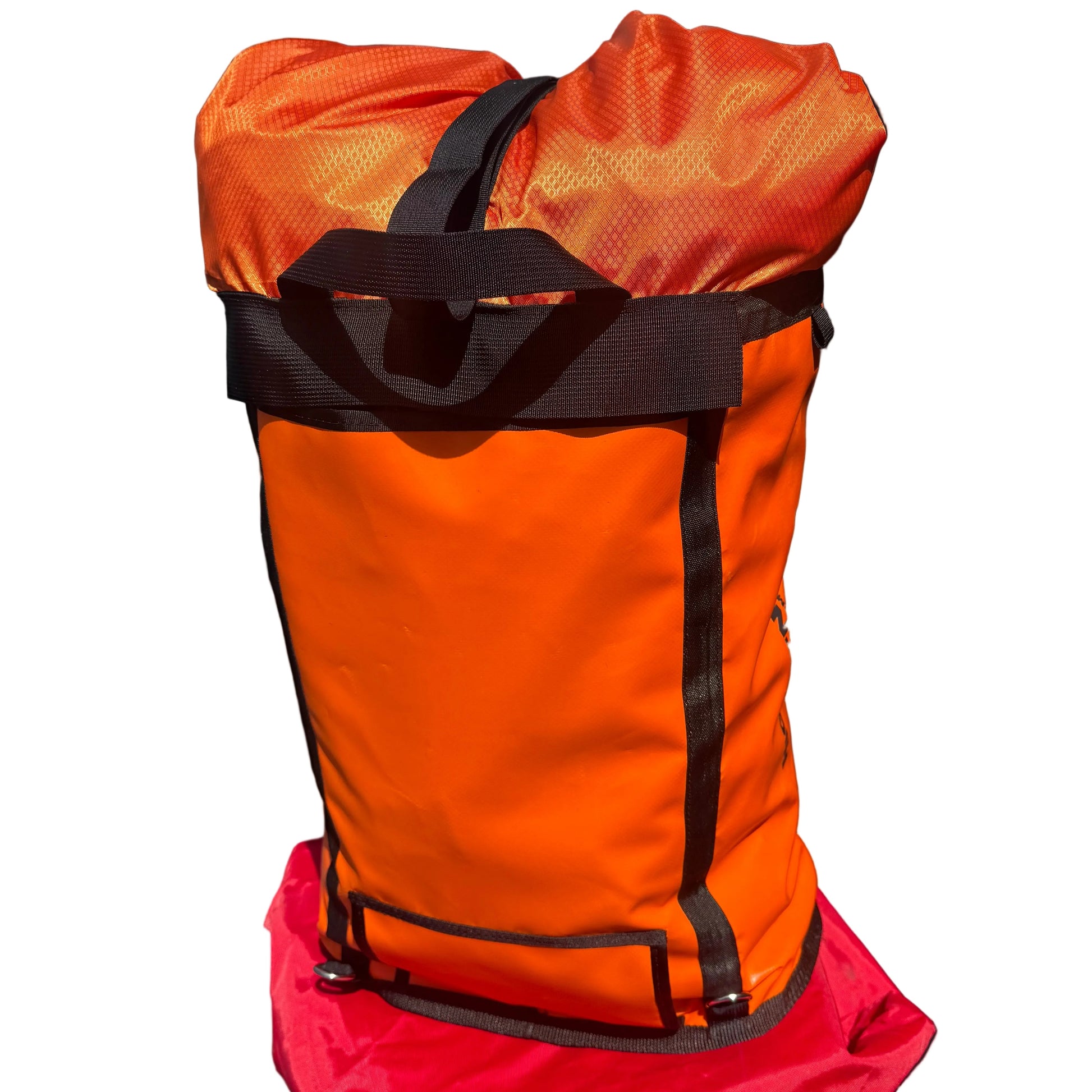 An orange waterproof stuff sack with black straps and trim, resting on top of a red bag against a dark brick wall.