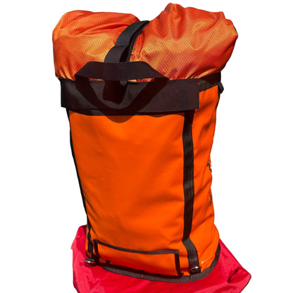 An orange waterproof stuff sack with black straps and trim, resting on top of a red bag against a dark brick wall.