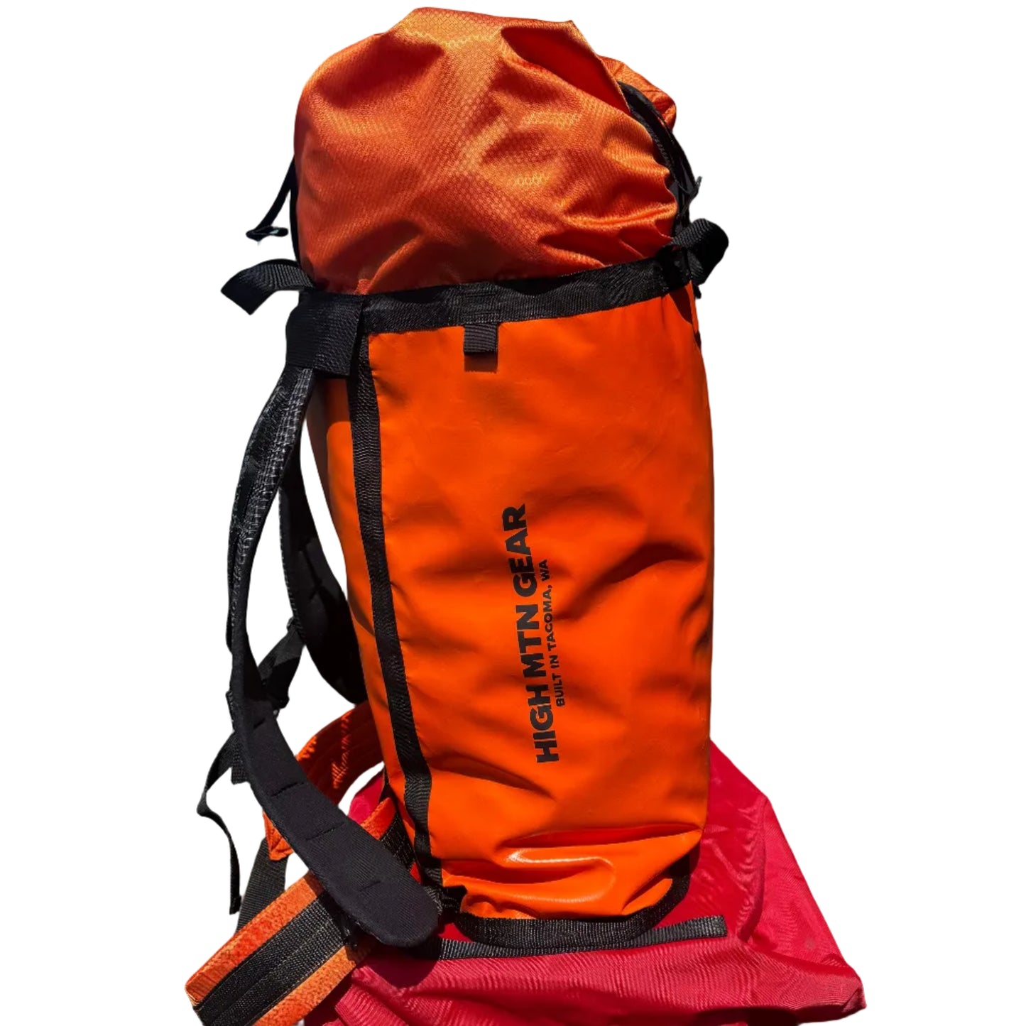 An orange High Altitude Gear dry bag or backpacking storage bag with black straps and buckles, standing upright outdoors against a dark gray brick wall and metal building corner.