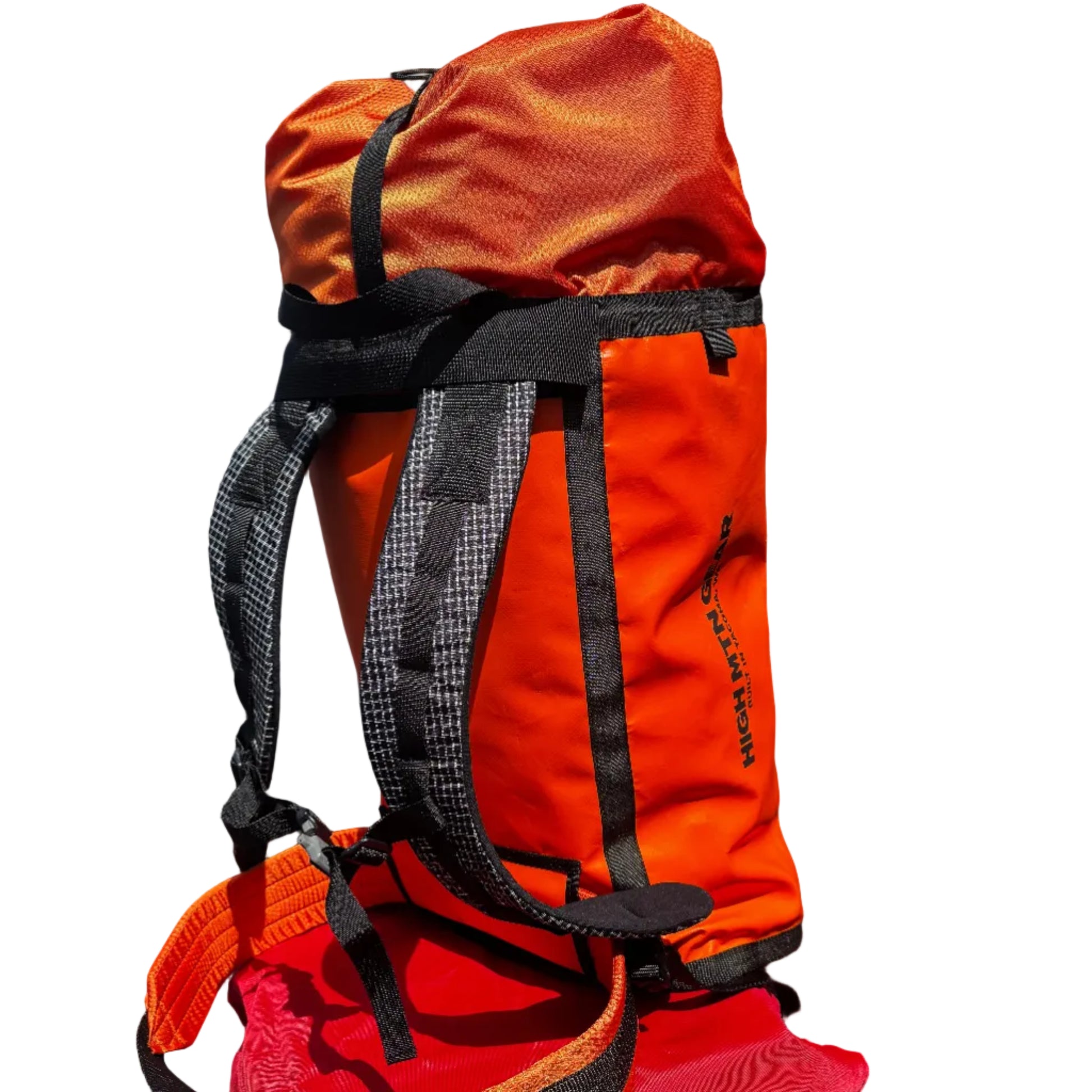 An orange waterproof backpacking bag with black straps and buckles, resting on top of a red bag against a dark gray brick wall.
